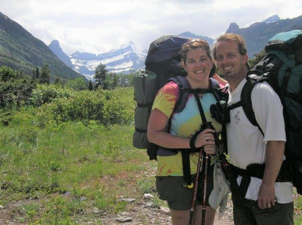 Amy and her husband wearing their backpacks on a trail in Glacier National Park with a view of the park's iconic glacier-carved mountain peaks in the distance.s, after crossing the river using the Cosley Lake Outlet Safety Cable.