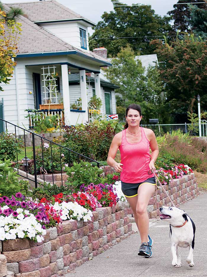 Woman jogging on an urban sidewalk with her leashed dog running beside her, looking up at her lovingly.