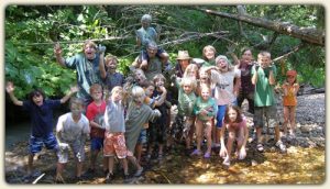 Group of youth summer campers enjoying the rugged outdoors at Twin Eagles Wilderness School. Kids wearing shorts, t-shirts, standing in the forest, one sitting on a tree branch.