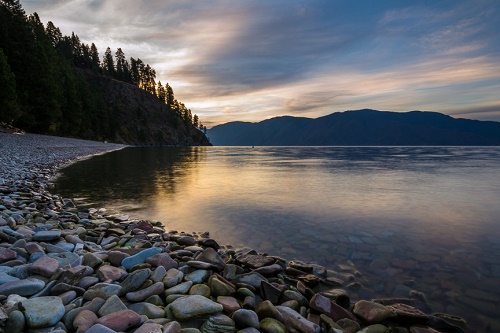 Still waters of Lake Pend Oreille at sunset, with faint orange reflection on the water and rocky shoreline and forested hills in the distance.