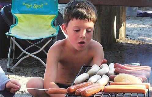 Young boy looking at the campfire grill covered in hotdogs and brats coving over the fire.