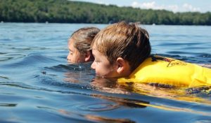 Two children swimming on their bellies in a lake, with chins in the water, while wearing life jackets.