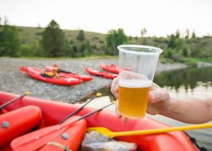 Hand holding a plastic cup of beer at the beech with inflatable rafts and kayaks on the rocky beach in the background.