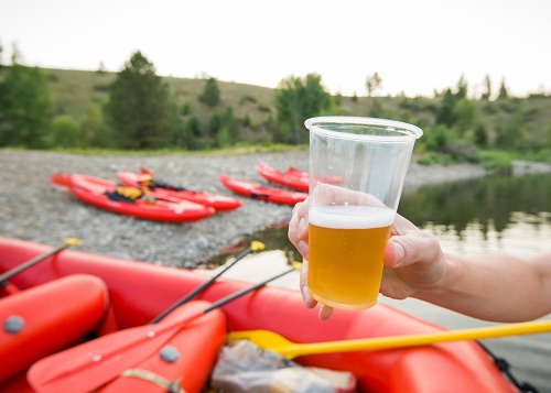 Hand holding a plastic cup of beer at the beech with inflatable rafts and kayaks on the rocky beach in the background.