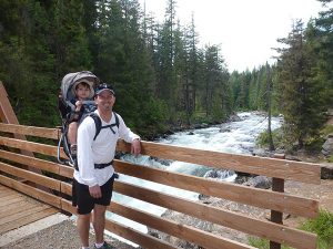 Dad with his toddler son in a backpack carrier -- standing on a bridge over the whitewater of Icicle Creek in the national forest near Leavenworth, Washington.