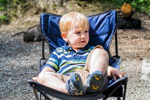 Child sitting in a camp chair at a campsite, with trees in the background.