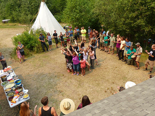 Looking down at Twin Eagles Wilderness School families, children and adults, standing on the dirt gathered in large circle with a smaller circle in the middle. White teepee and trees in the background.
