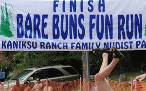 Large Finish Line banner that reads, "FINISH BARE BUNS FUN RUN - Kaniksu Ranch Family Nudist Park," with a view of people's heads and someone's legs and shoed-feet as if doing a handstand.