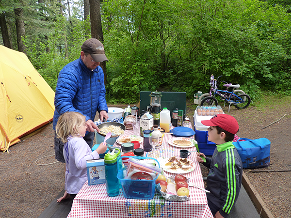 A dad serving breakfast to kids at the family campsite picnic table.
