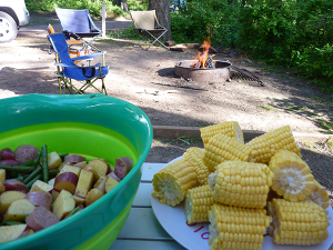 Bowl of cut vegetables and raw shrimp, and plate of corn cobs on a picnic table at a campsite.