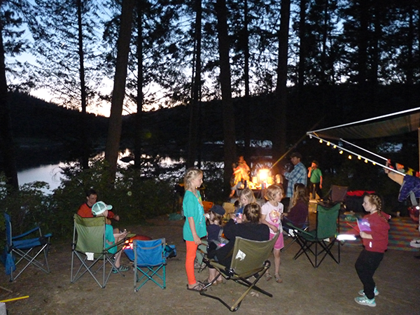 Family camping with children and adults gathered around a campfire eating hotdogs.