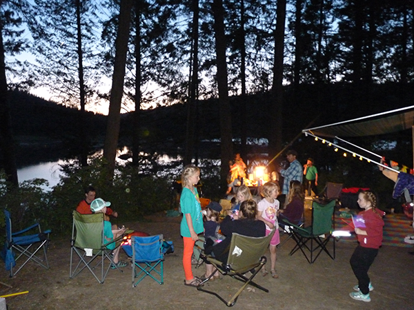 Campsite with parents and children sitting in camp chairs around campfire eating, with a pop-up tent trailer with awning and picnic table with lantern. View of the lake in the background.