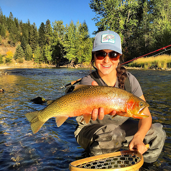 Woman holding a rainbow trout with one hand, with her fly fishing net in her other hand, while wading in the North Fork of the Coeur d'Alene River.