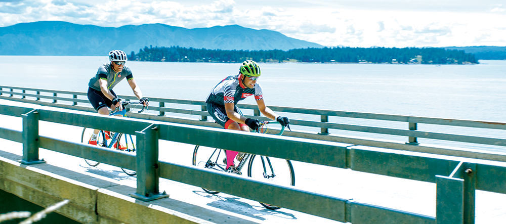 Two male racers cycling on Long Bridge across Lake Pend Oreille, with mountains in the distance, during the Chafe 150 event in Sandpoint, Idaho.