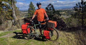Hank Greer with bike wearing a neon orange shirt with neon orange travel packs attached to his bike.