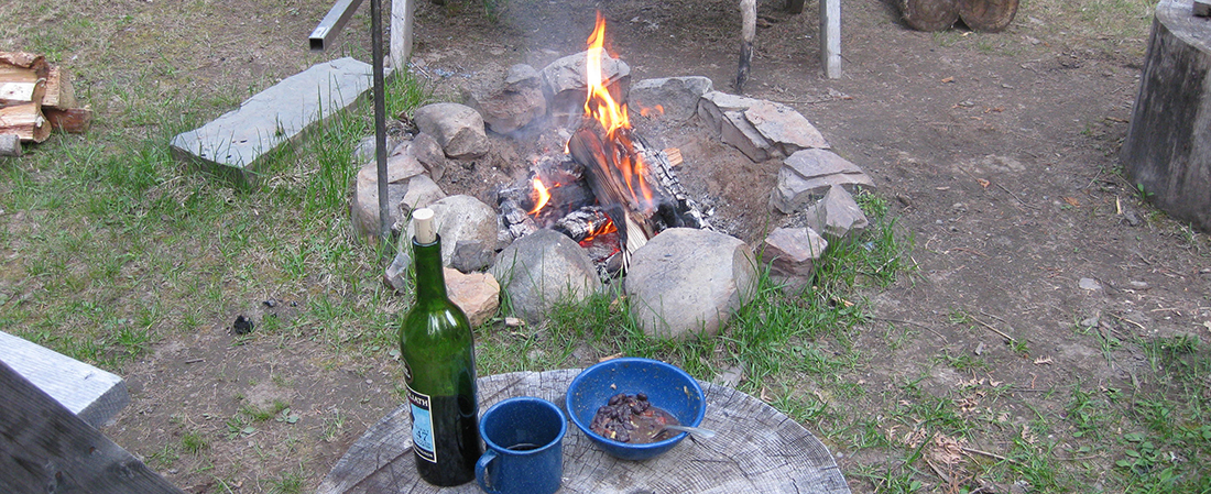 Photo of campfire with bottle of wine, coffee cup, and bowl resting on a tree stump.