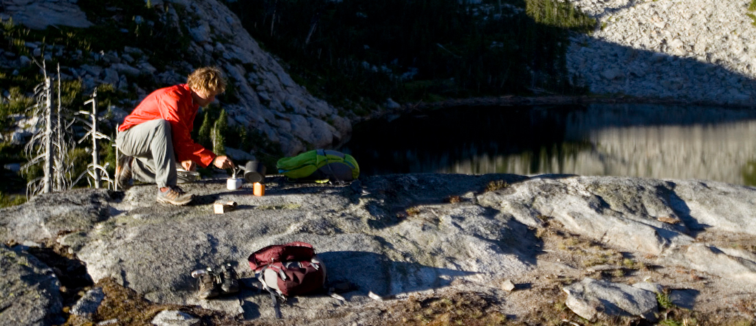 An adult woman lying on a camp pad journaling and an adult man boiling water during a backcountry camping trip in the Selkirk Mountains of Northern Idaho.