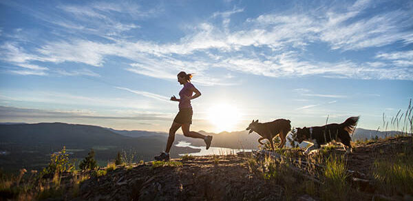 An Adult Woman Running With Two Dogs On Top Of A Mountain Overlooking The Pend Oreille River In Sandpoint, Idaho