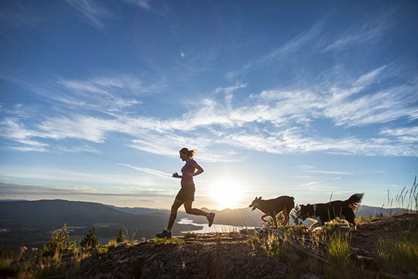 An Adult Woman Running With Two Dogs On Top Of A Mountain Overlooking The Pend Oreille River In Sandpoint, Idaho