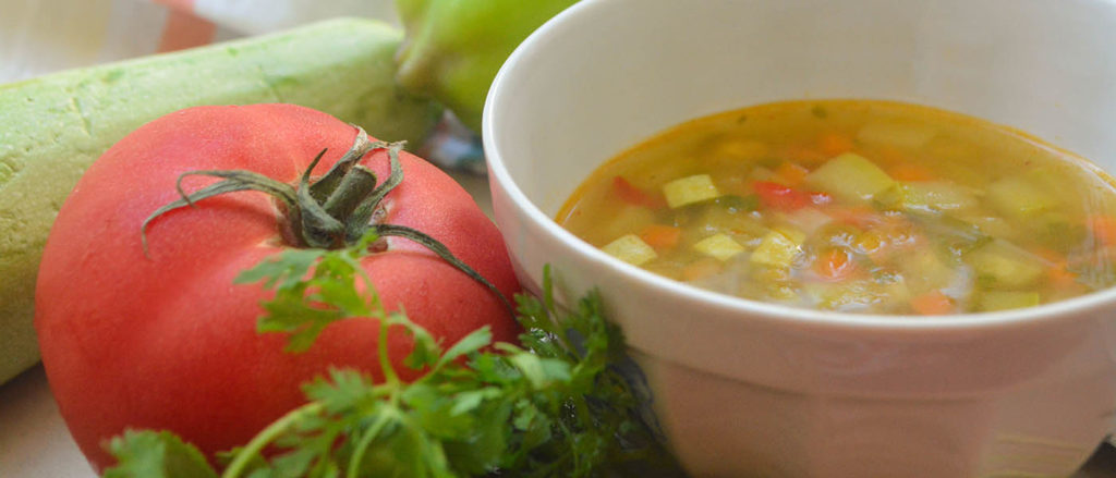 Photo of vegetable soup in bowl next to tomato and parsley.