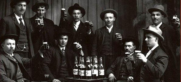 Black and white vintage photo of men in black suits standing next to bottles of beer.