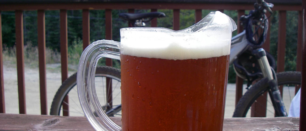 Photo of beer on picnic table with bike in the background leaning against deck railing.