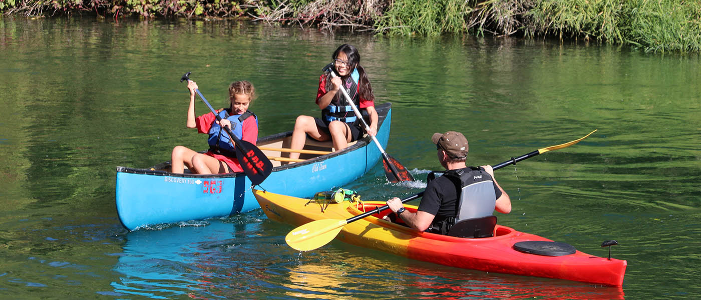 Two teenage girls paddling a blue canoe and a man paddling a red and yellow kayak on the Little Spokane River at Saint George's School.