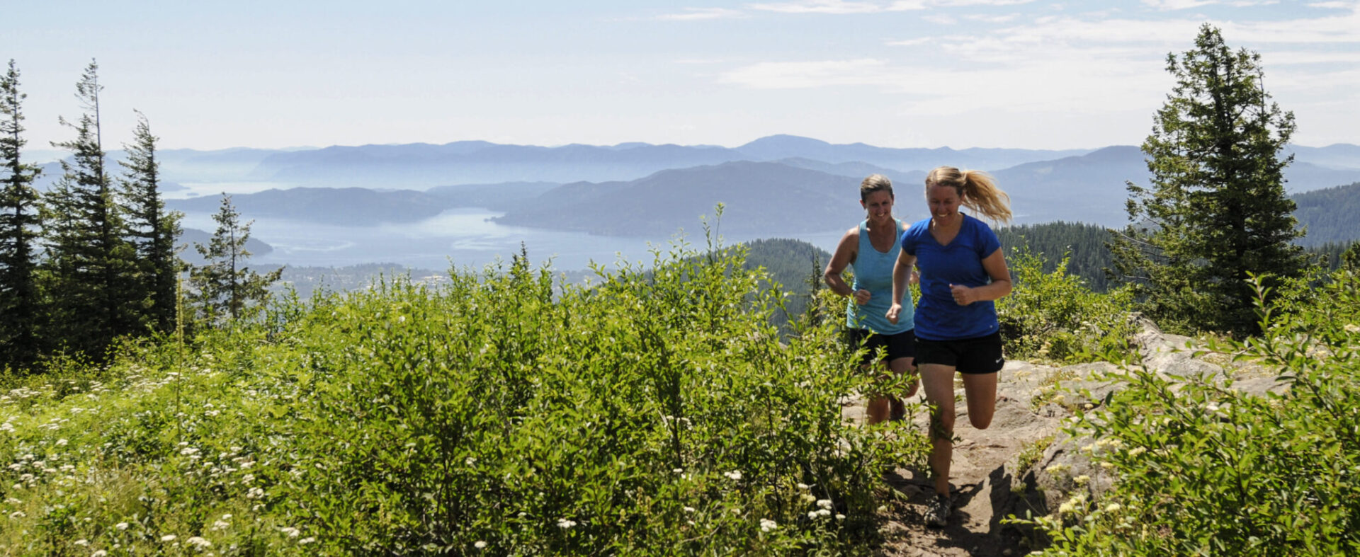 Two runners racing along a dirt trail on Schweitzer Mountain with a view of Lake Pend Oreille in the far distance.