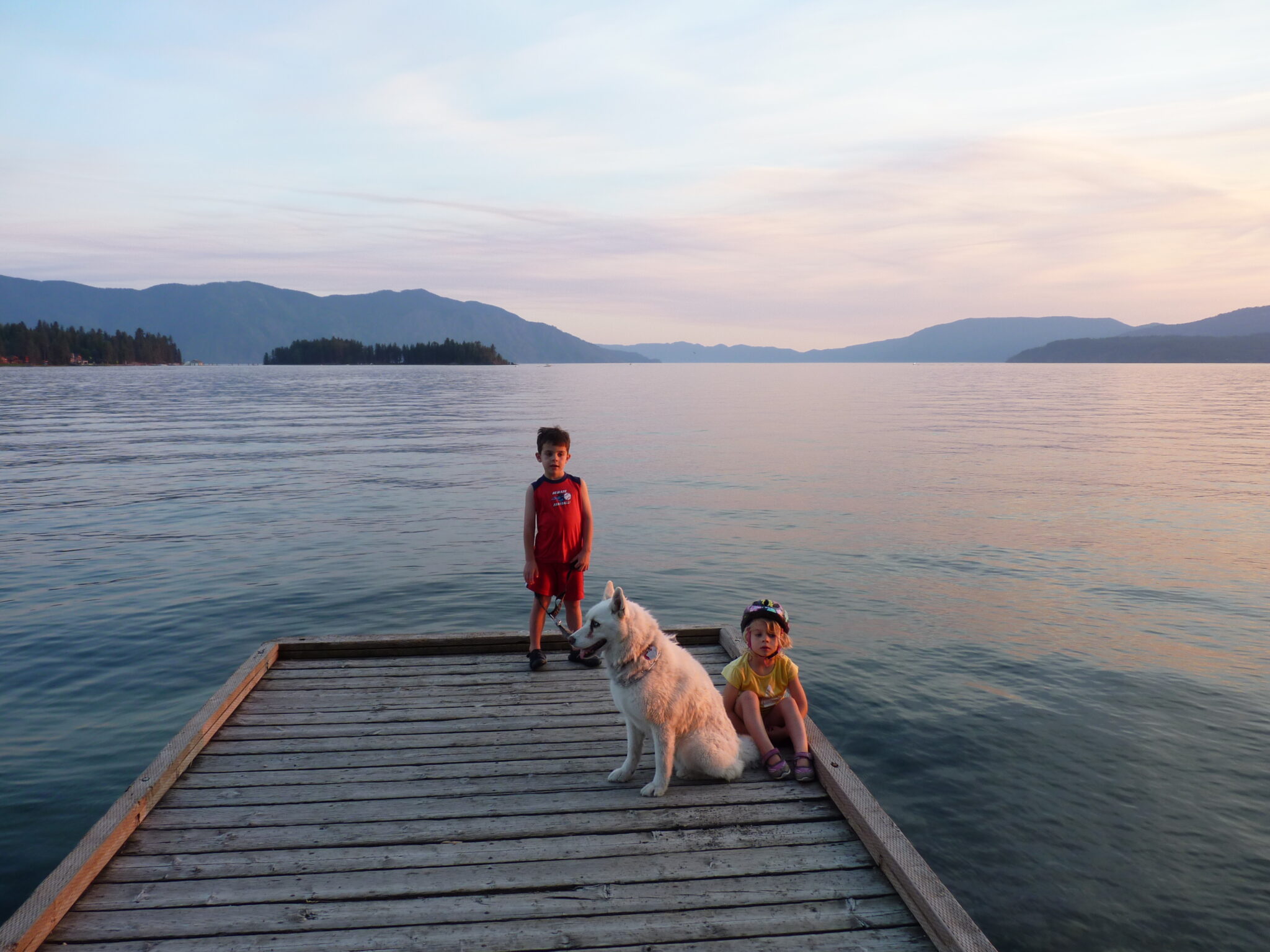Two young children with their dock standing on a dock at sunset at Lake Pend Oreille, with forested hills in the far distance.