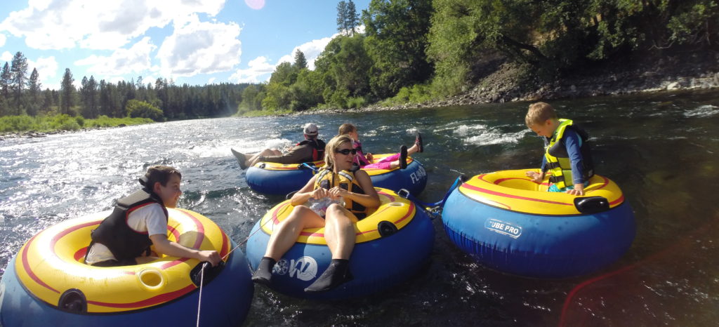 Mom, dad, and children tubing on the Spokane River, using big blue and yellow inflatable tube rentals from FLOW Adventures. Small whitewater rapids on the water and trees along the riverbank.