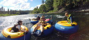 Mom, dad, and children tubing on the Spokane River, using big blue and yellow inflatable tube rentals from FLOW Adventures. Small whitewater rapids on the water and trees along the riverbank.