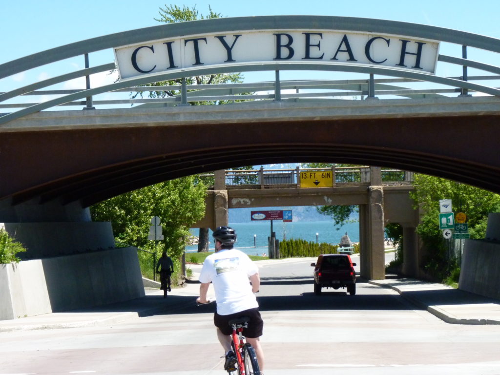 Man biking to City Beach in Sandpoint, approaching a big overhead sign at the park entrance.