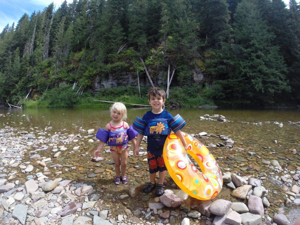 Two young children wearing personal flotation devices and holding a tube while standing on the rocks of the North Fork of the Coeur d'Alene River.