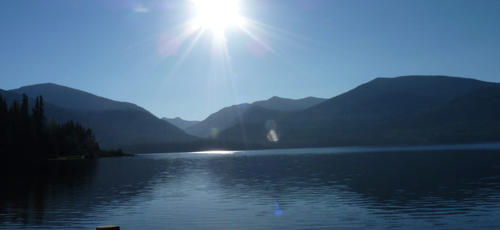 Still water of Priest Lake with mountain peaks and sun in the background.