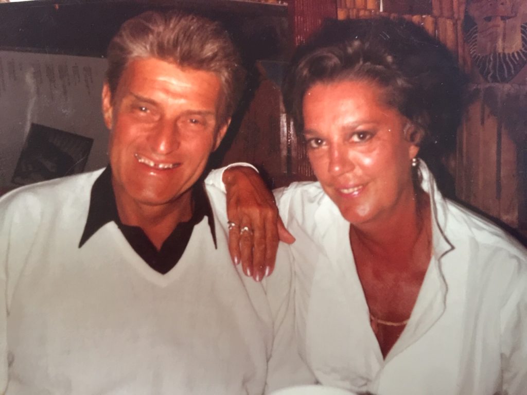 George and Lois Hill smiling at the camera in a circa 1970s photo. George is wearing a wide-collared black button-up shirt under a white V-neck sweater and Lois is wearing a white deep-V-neck blouse accessorized with goal necklace. Her right hand and arm is resting atop George's left shoulder.