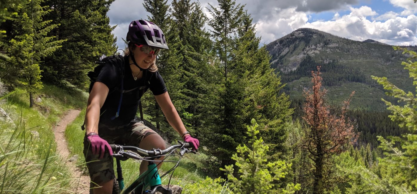 Woman mountain biking on a dirt trail with a view of trees and mountain peaks in the background.