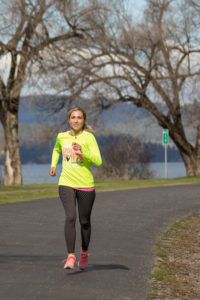 Woman running on the Centennial Trail in Spokane, with a few of trees and the Spokane River in the background.