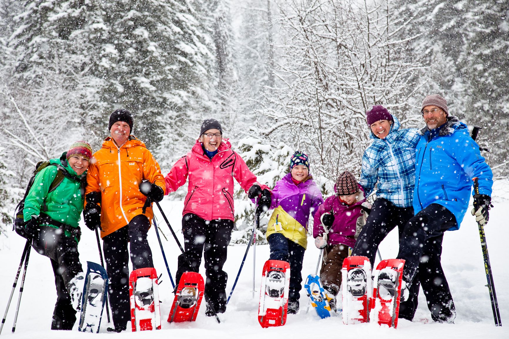 Group of 6 people, adults and children, each wearing a bright different-colored coat, smiling at camera and showing their snowshoes with one foot raised off the snow.