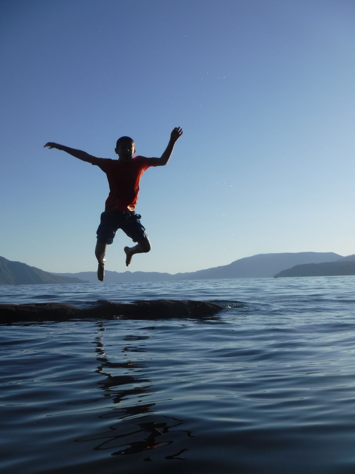 A boy jumping off a log floating in Lake Pend Oreille, with a view of hills in the far background.