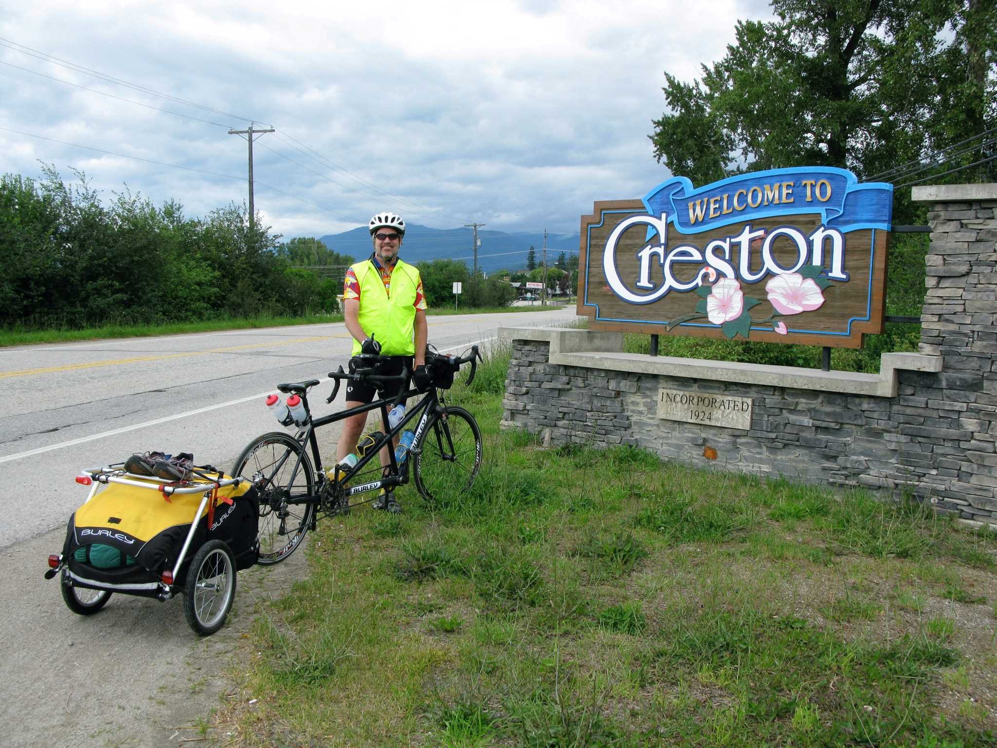 A bike rider taking a break with his gear in front of a town sign.