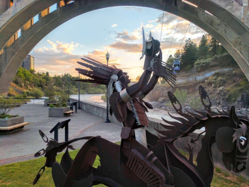 Metal sculture of Spokane Tribal member sitting atop horse and holding up a salmon. Huntington Park in downtown Spokane, with Spokane River alongside and Monroe Street Bridge above.