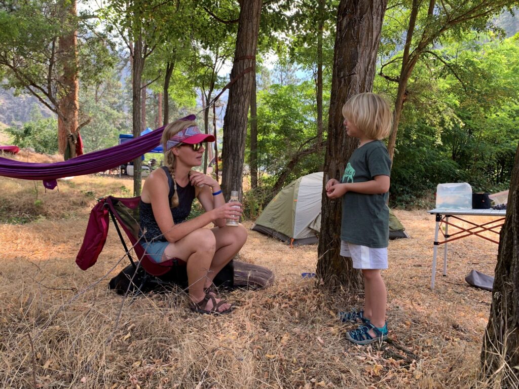Treed campsite with a mom in a camp chair and child standing, and tents in the background.