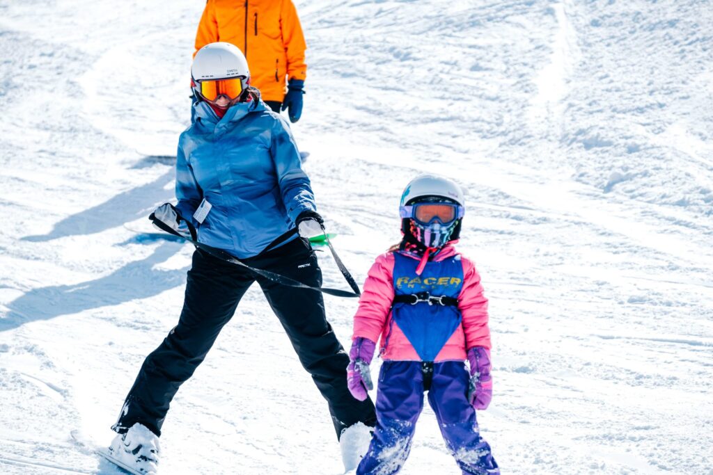 Young girl learning to ski with harness and safety reins held by instructor skiing behind her to help child make turns and control speed.