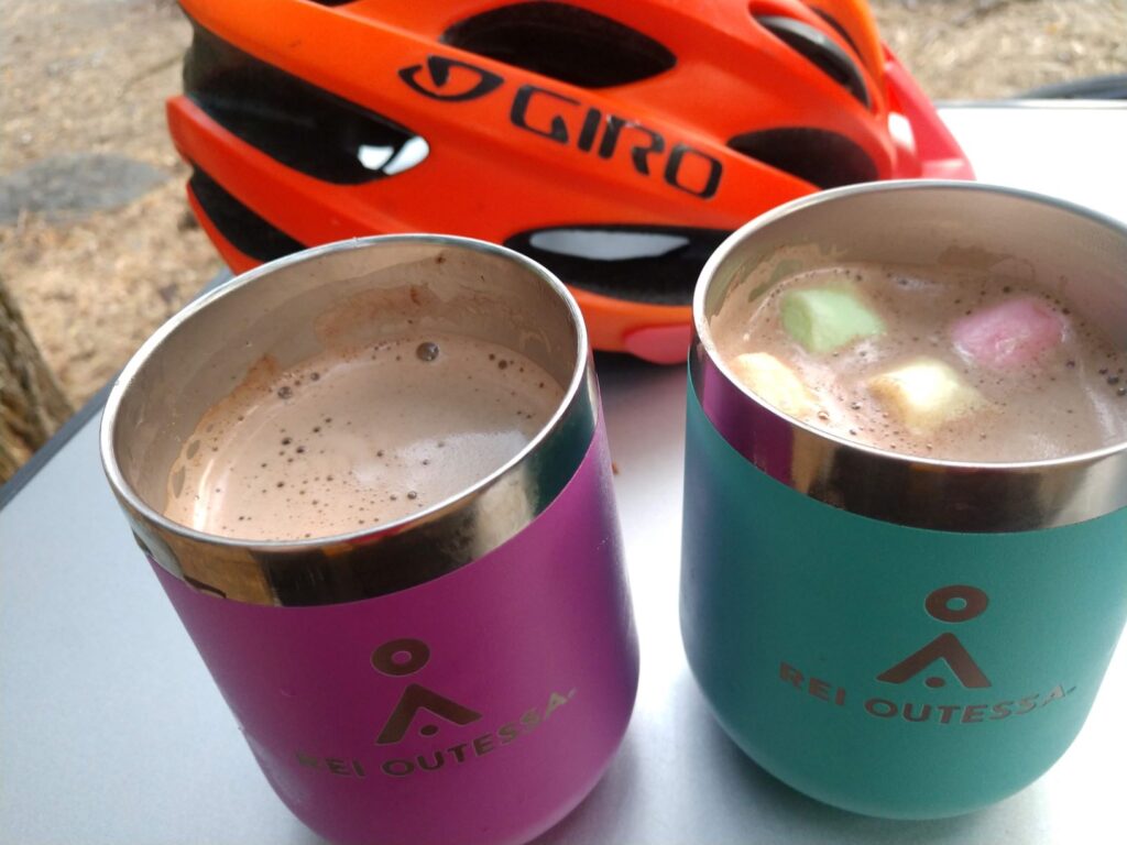 A table with two stainless steel mugs filled with hot cocoa, topped with marshmallows, and a bike helmet behind them on the tabletop.