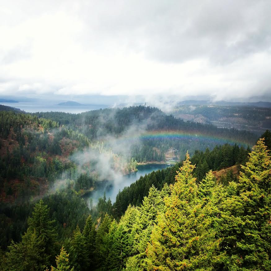 View from Timberline Adventures zipline course of of Beauty Bay at Lake Coeur d'Alene, featuring forested hillsides, wispy clouds, and a rainbow.