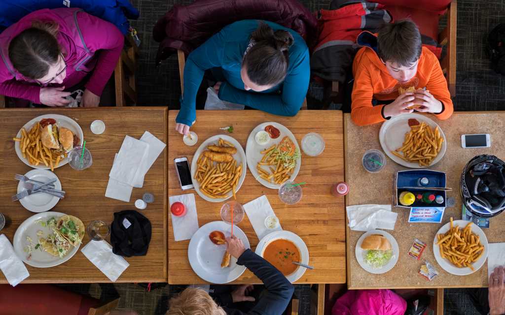Looking down at a ski lodge rectangle table with plates of food and cups, and skiers sitting along each side.