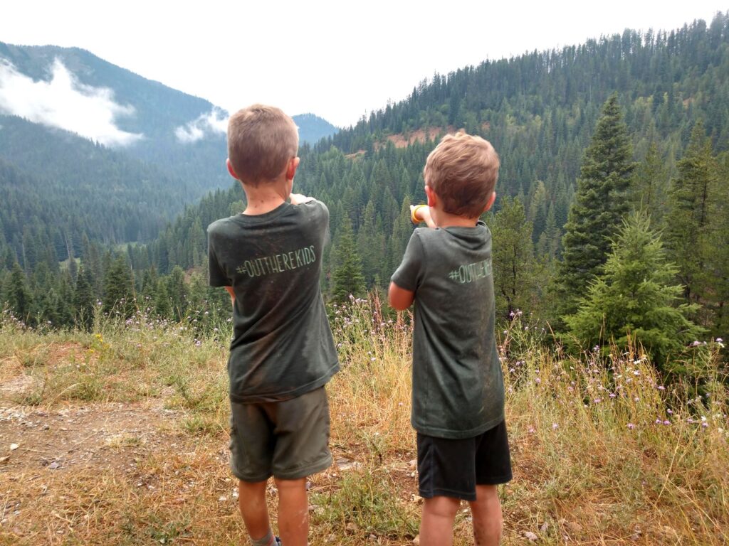 Two children wearing t-shirts that say #OutThereKids on the back, standing in a clearing and looking and pointing to the view of a forested hillside in the distance.