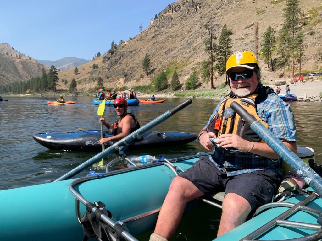 Man paddling an inflatable raft on the Salmon River, with kayakers in the backyard, and dry, rugged hilly landscape along the river.