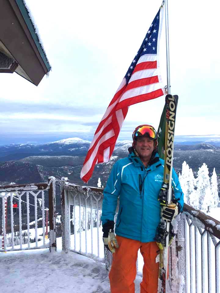 Fred Nowland in his ski gear on a resort deck with the mountain view and American flag behind him.