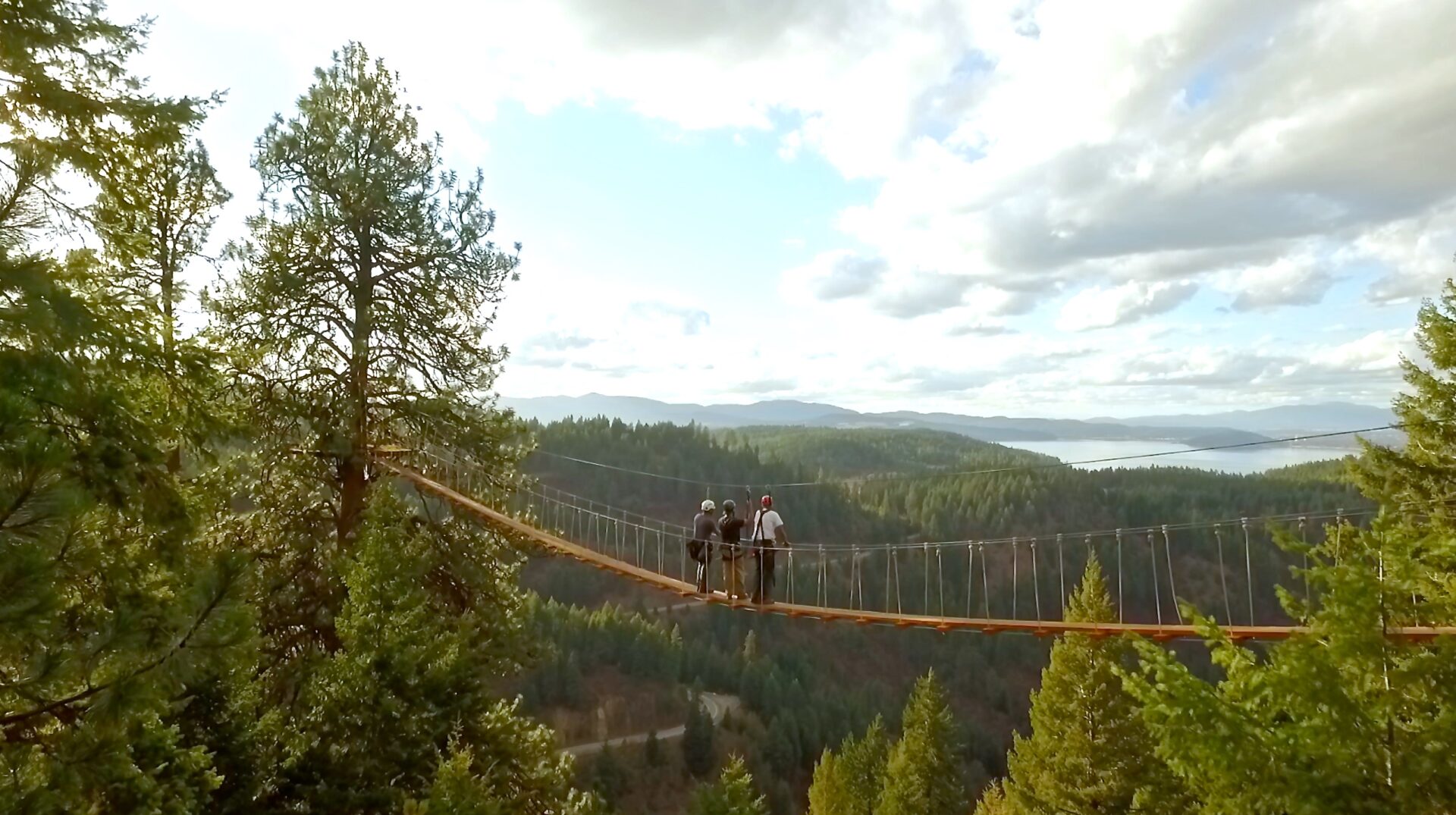 Two people standing on a high suspension bridge overlooking forested hillside with Lake Coeur d'Alene in the far distance.